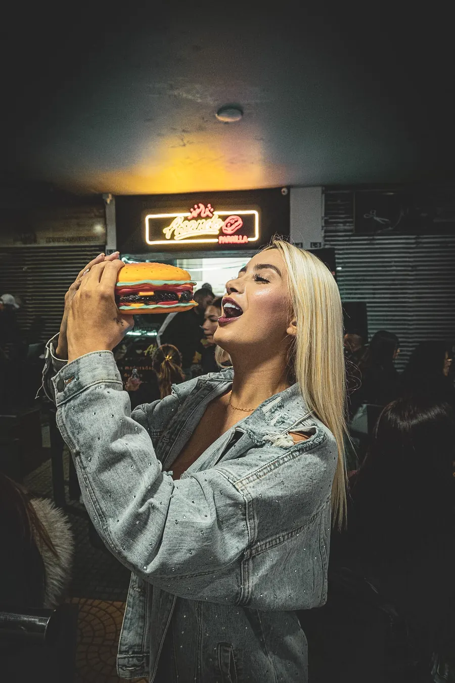 Clienta haciendo como que muerde una hamburguesa frente al letrero de neón de Asando Parrilla