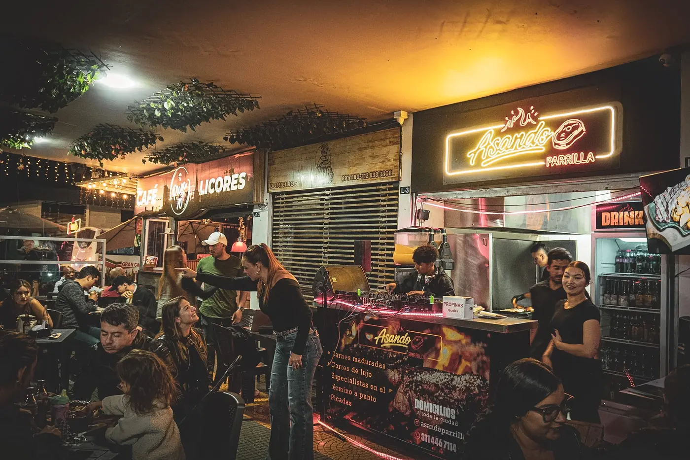 Asando Parrilla de noche: letrero de neón, la parrilla, el equipo atendiendo, un DJ y las mesas llenas